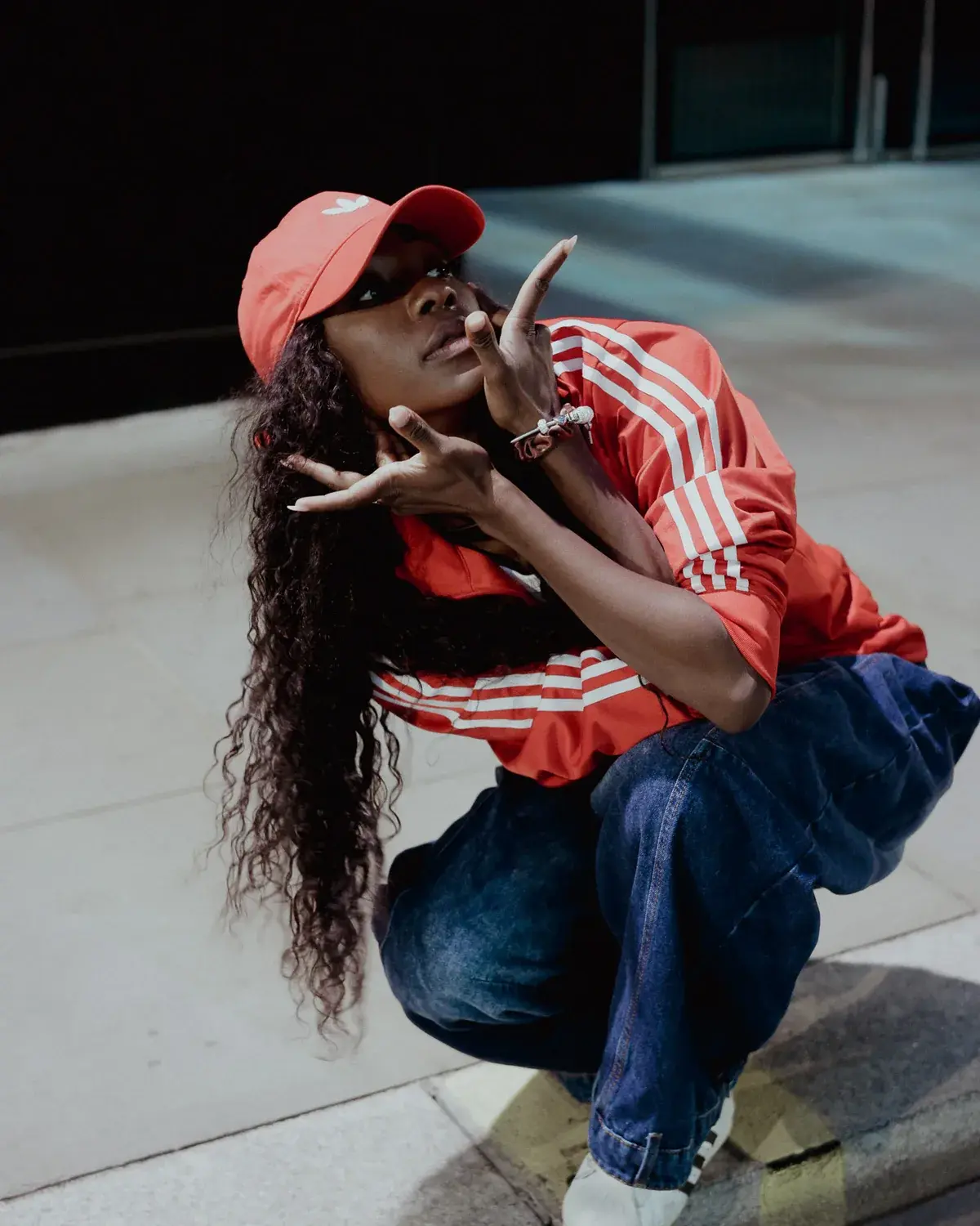 Cori D Lionne crouched low on a kerb at night, hands framed in front of her, in a red track top and cap.