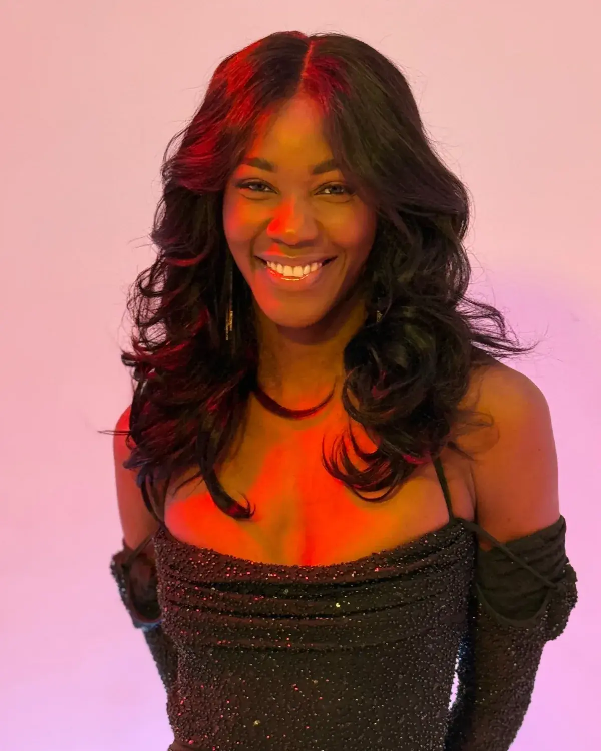 Cori D Lionne smiling under warm pink and orange light, hair loose over her shoulders.