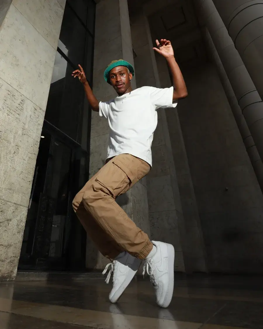 Tebza Diphehlo mid-jump outside a stone building, arms raised, wearing a green bucket hat and white T-shirt.