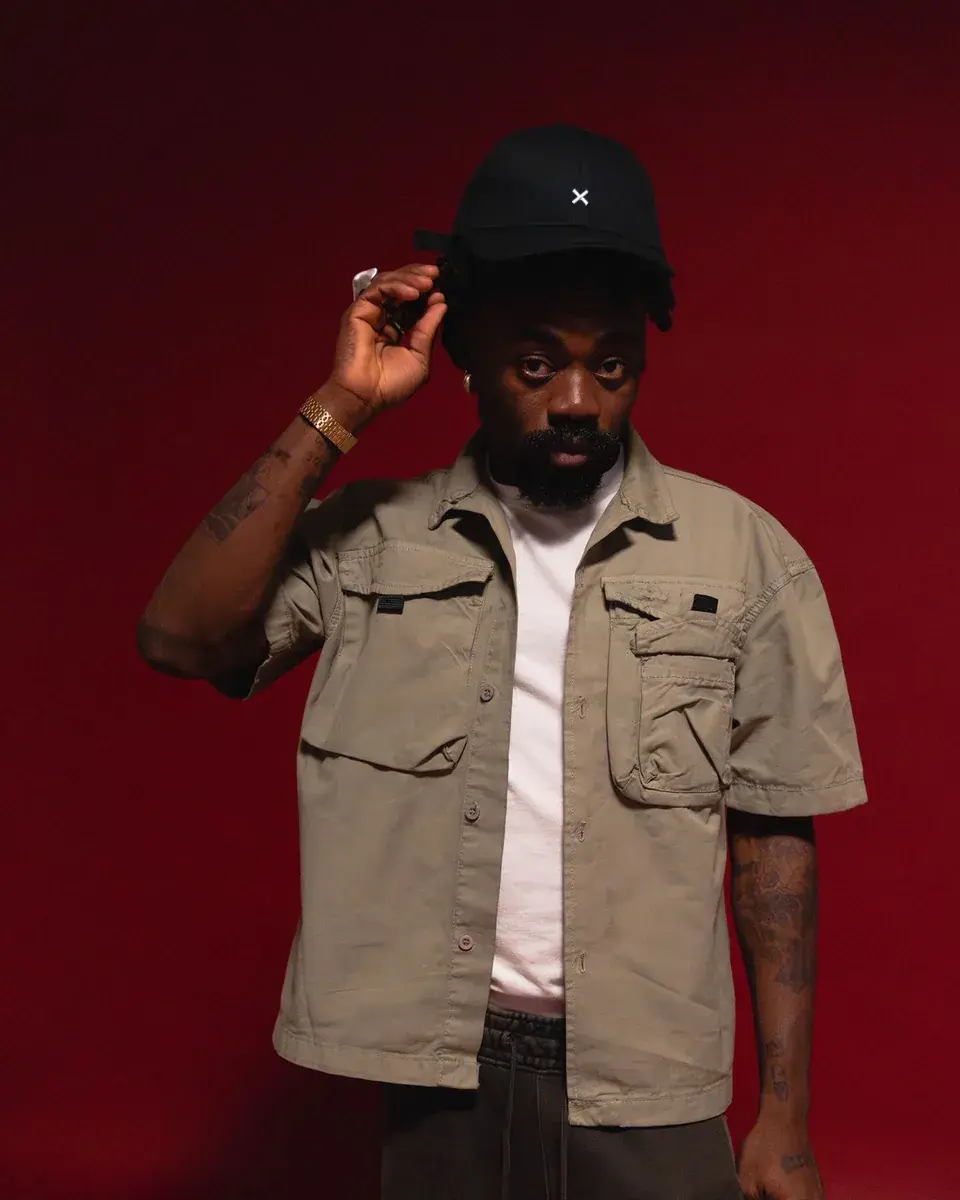 Yemi Osokoya standing against a deep red backdrop in a khaki utility shirt, one hand adjusting his black cap, looking straight to camera.