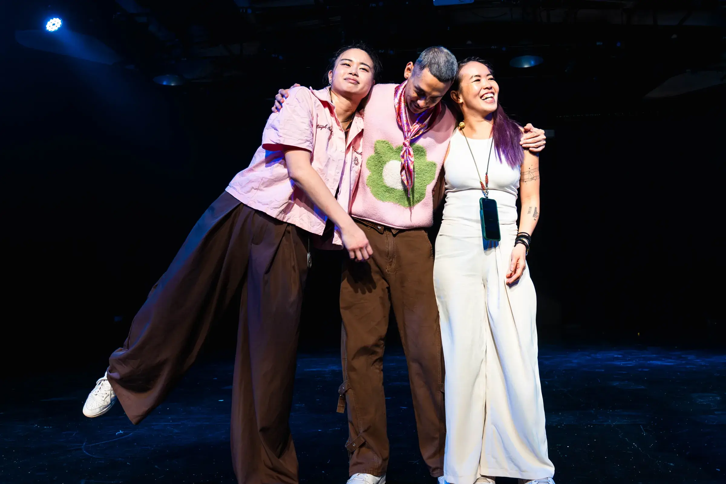 Three people lean into each other laughing, arms around shoulders, on a dark stage.
