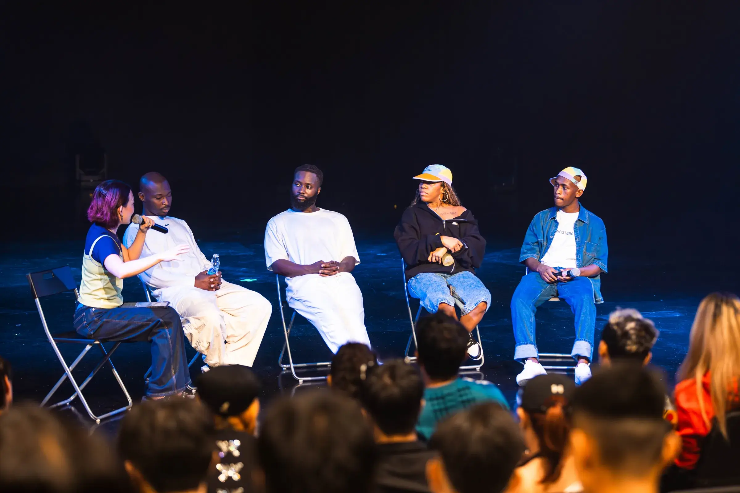 Five people seated in a row of chairs on stage for a panel discussion, audience heads in the foreground.