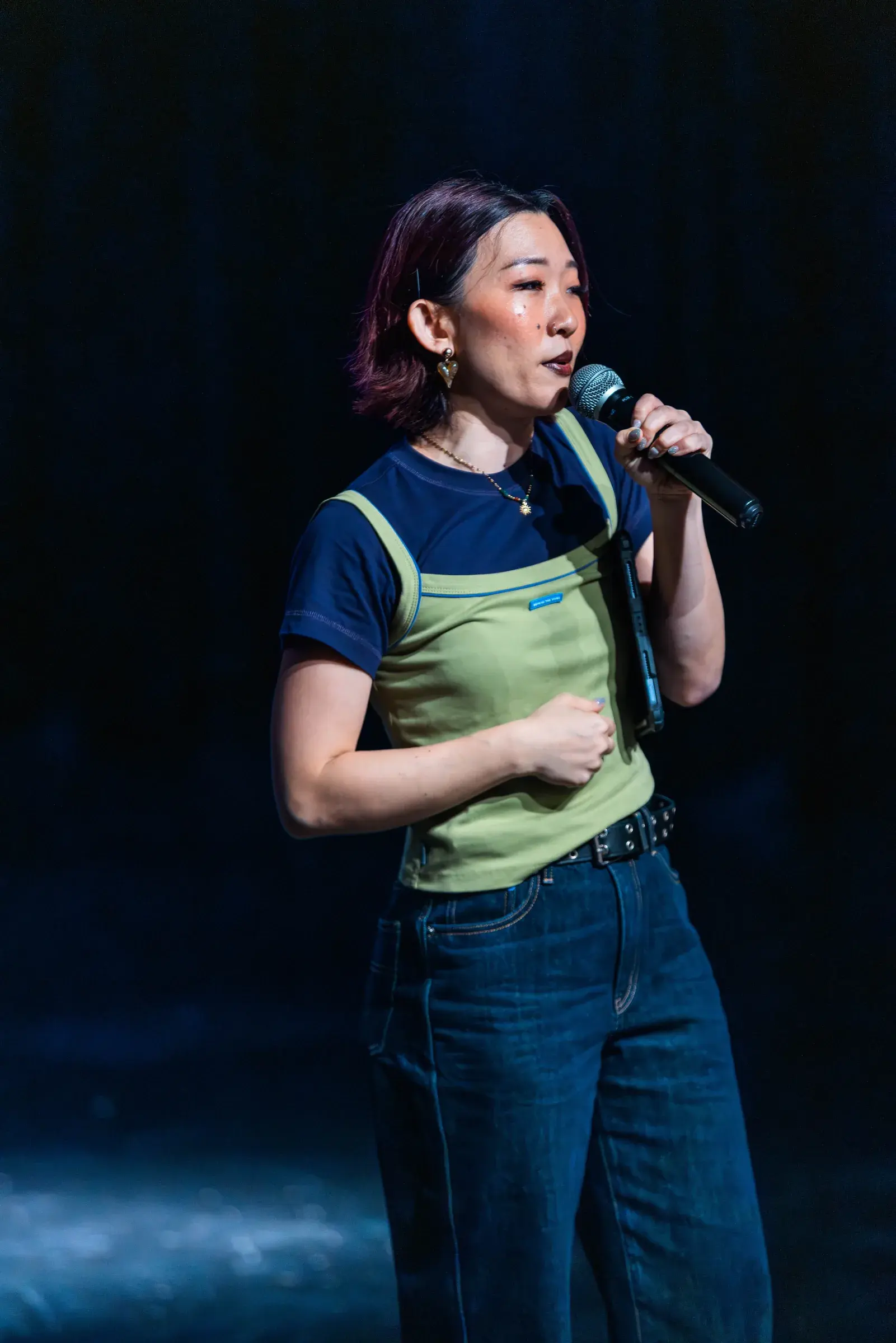 The host stands alone in a spotlight on a dark stage, speaking into a handheld microphone.