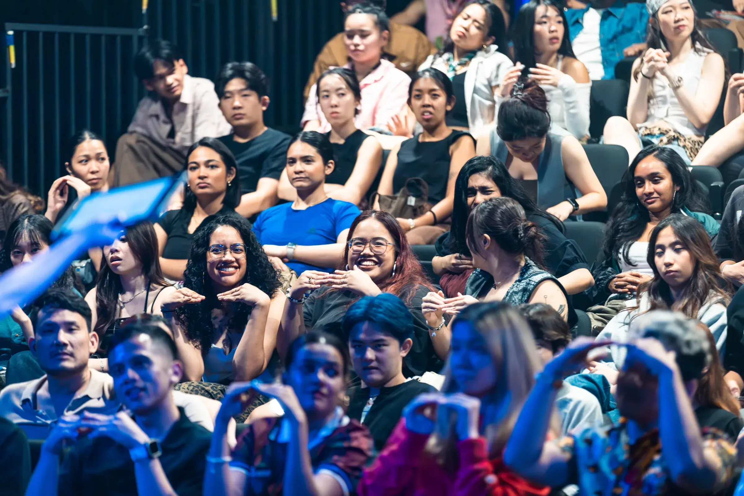 A packed audience in raked theatre seating, hands lifted above their heads as they watch the stage.