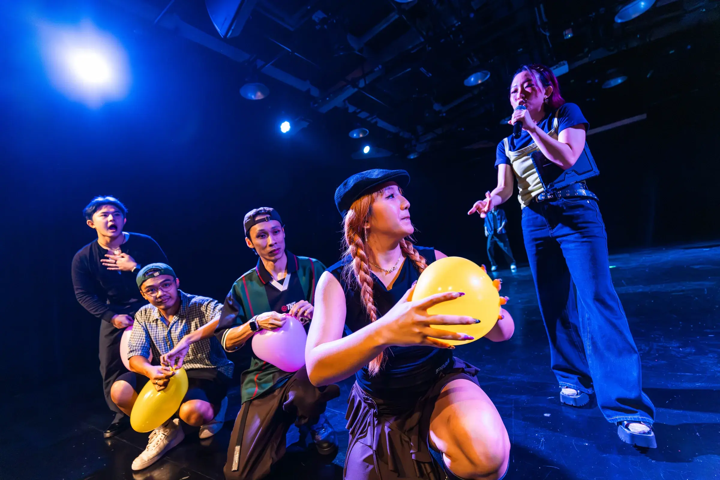 Performers kneel in a rough arc on stage passing and inflating coloured balloons as a host looks on.