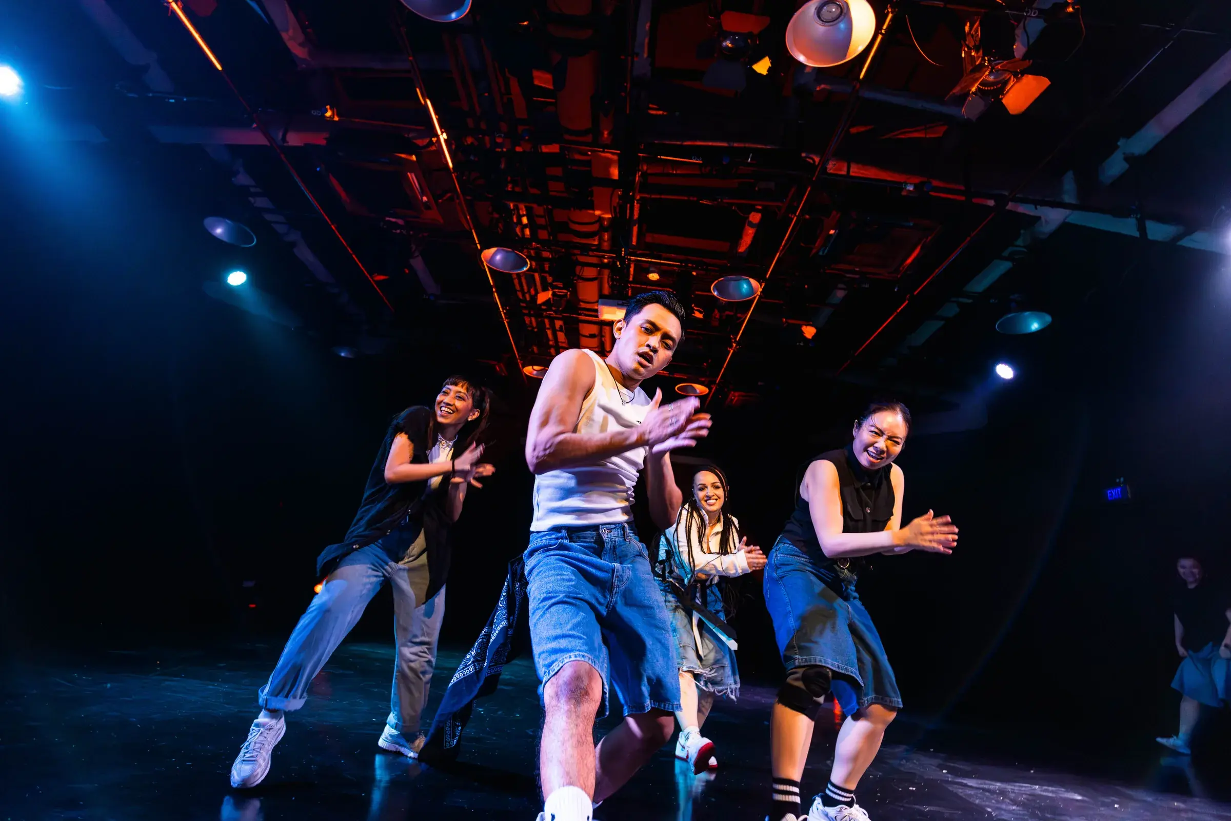 A dancer in a white tank top leans forward with arms extended, three dancers laughing behind him.