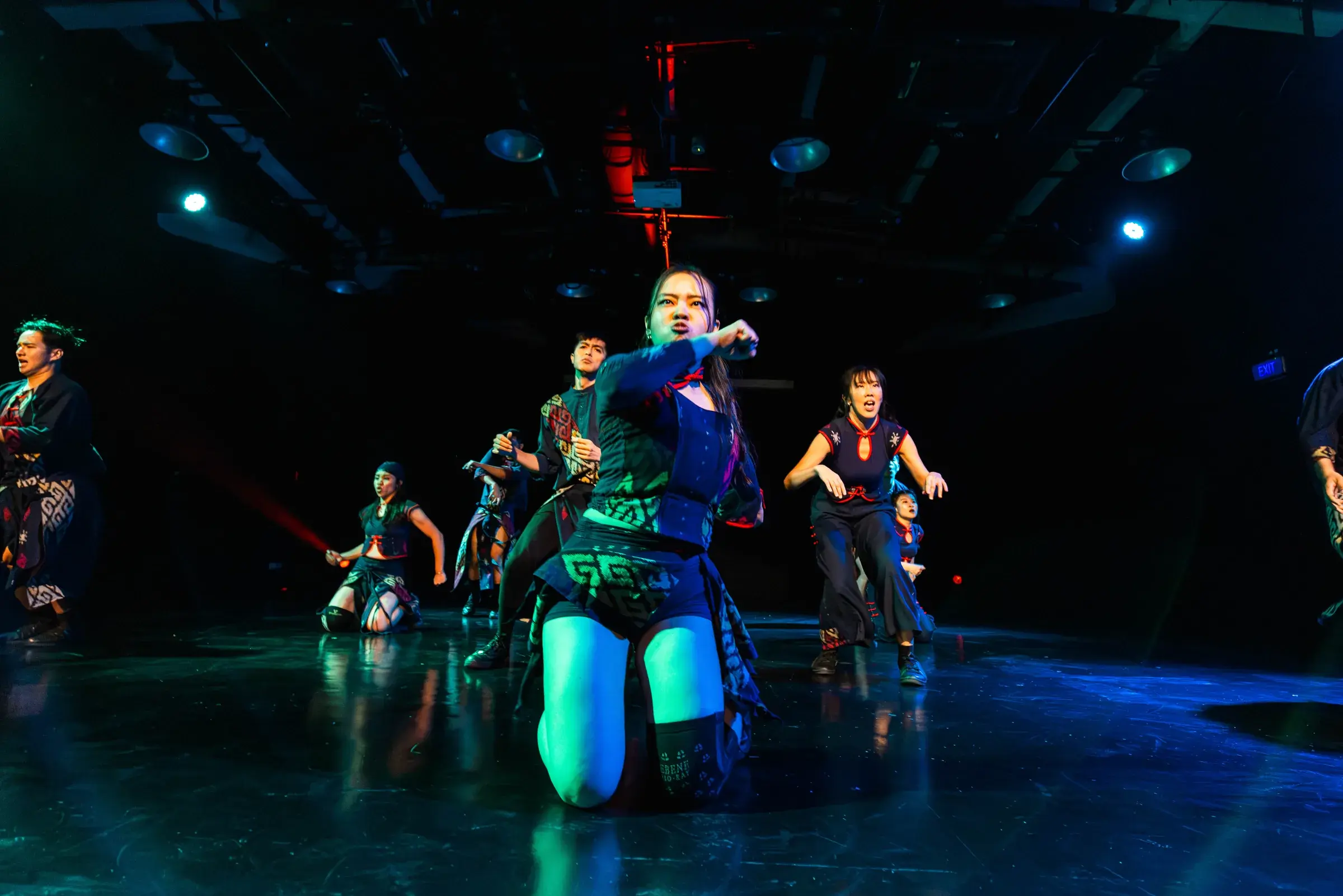 A dancer kneels centre stage with one arm raised, lit green, her crew fanned out behind.