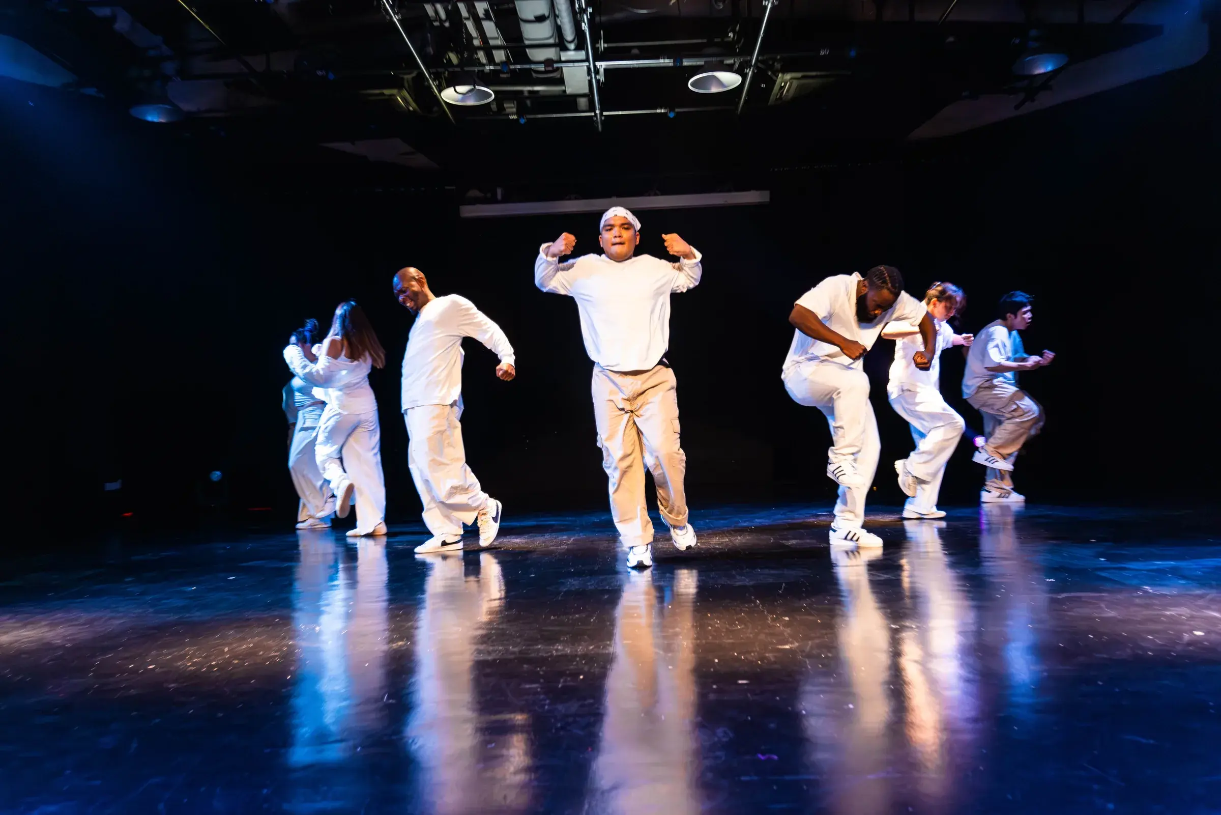 A dancer in cream flexes both arms overhead while the rest of the crew moves around him.