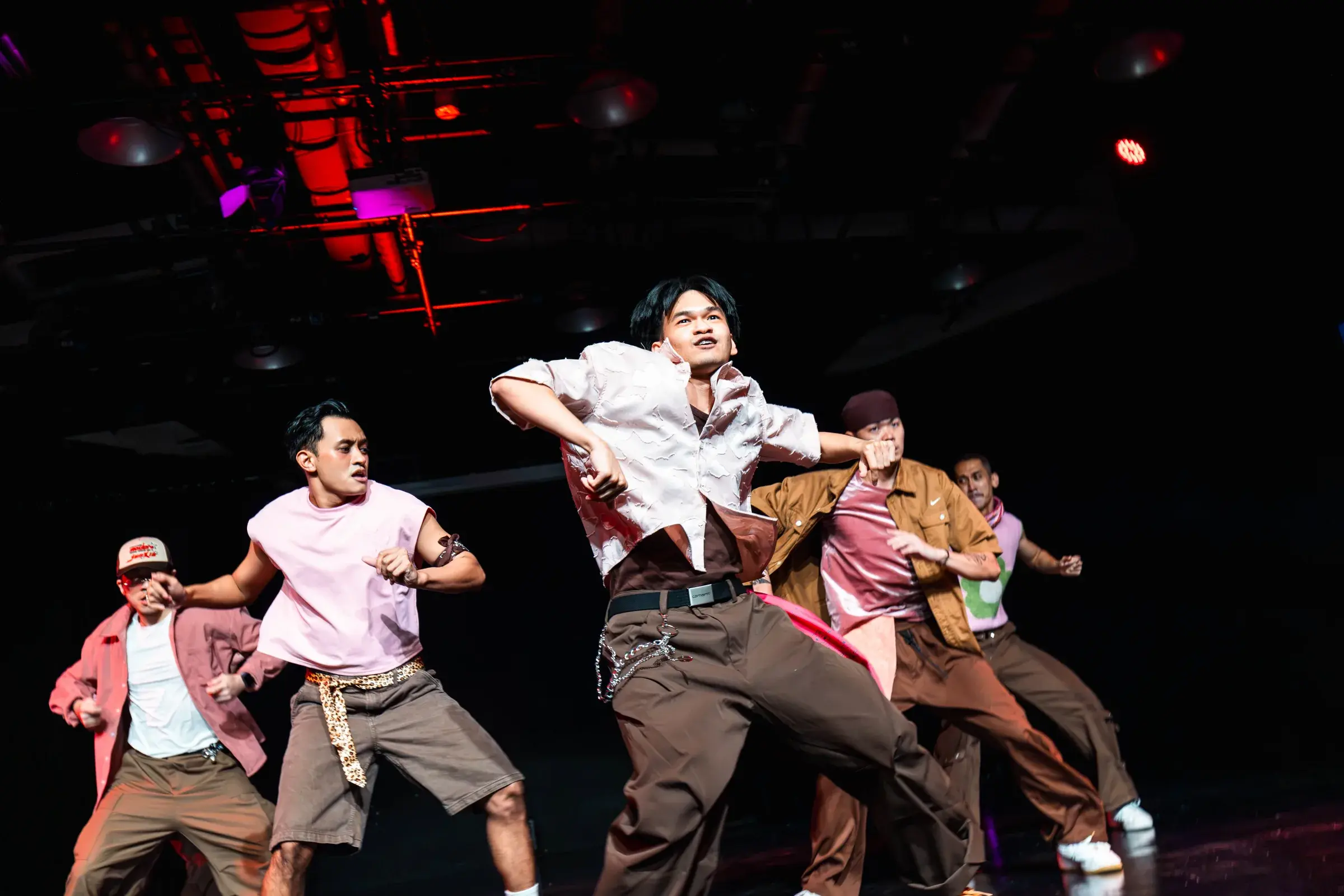 Five dancers in pink and brown cluster tightly together, the front dancer shouting with his arm up.