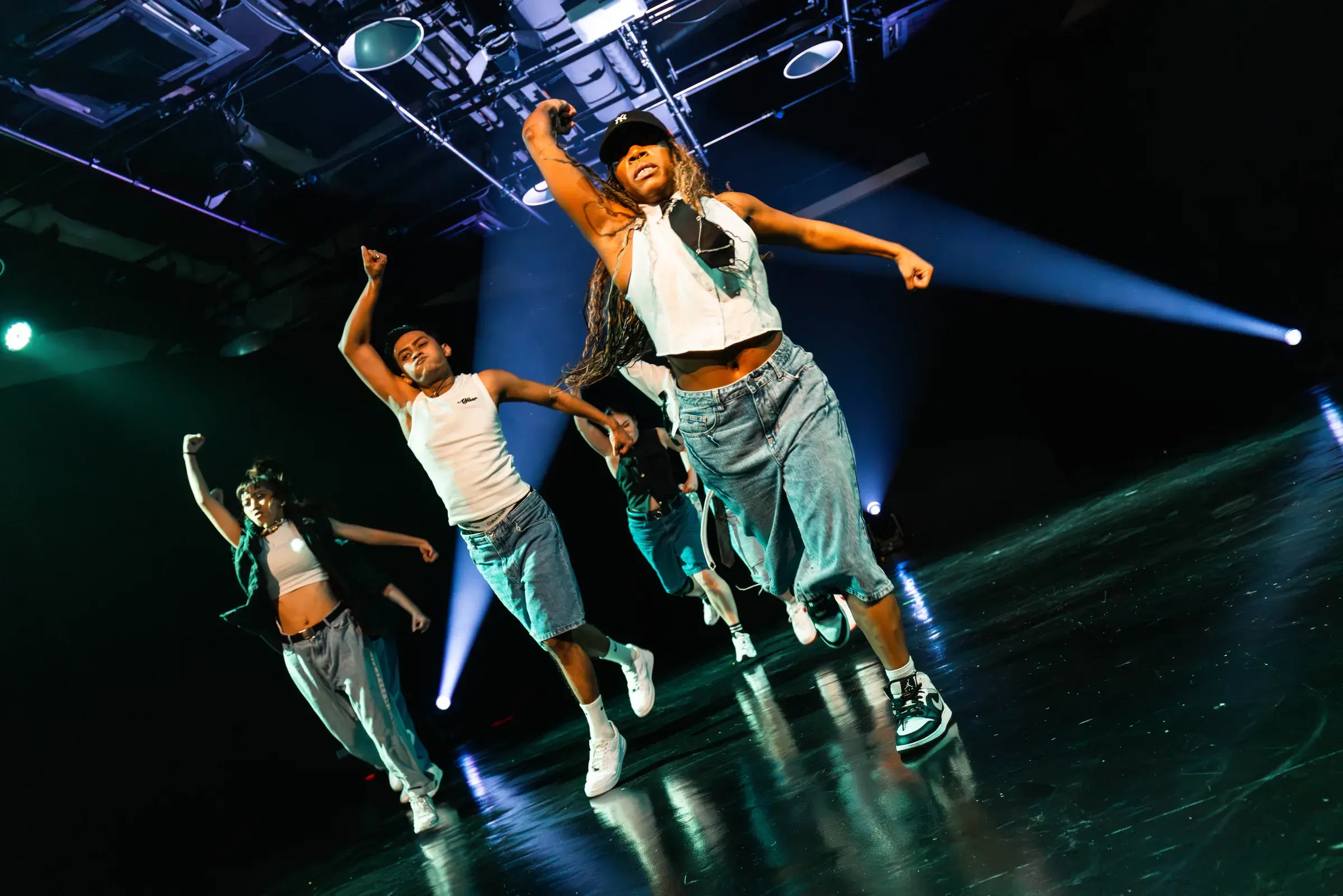 A dancer leaps into the air with arms raised as two crossed spotlight beams cut across the stage.
