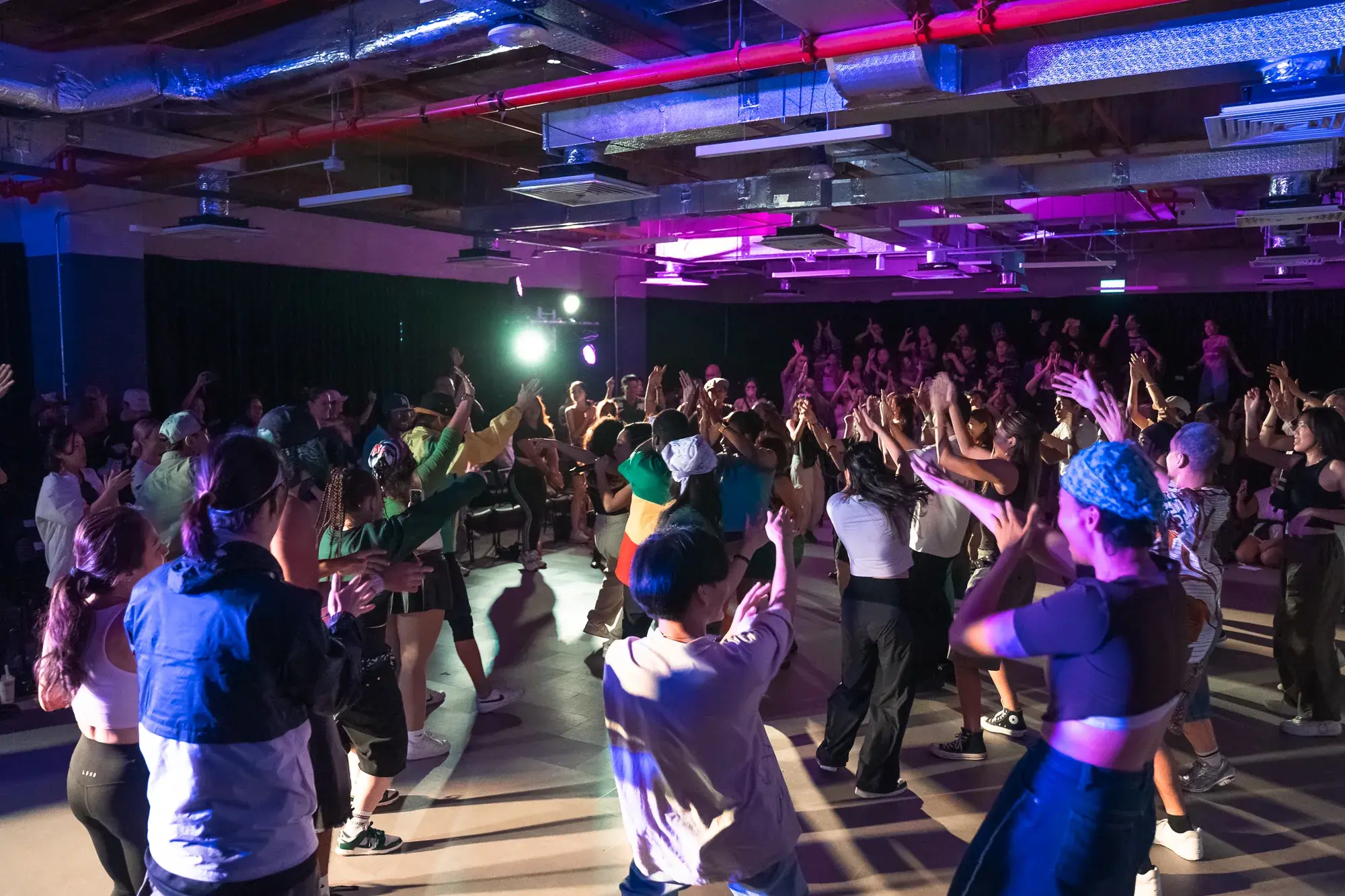 A packed studio at ACE 2025, dozens of dancers with their arms raised mid-routine under purple and blue lights.