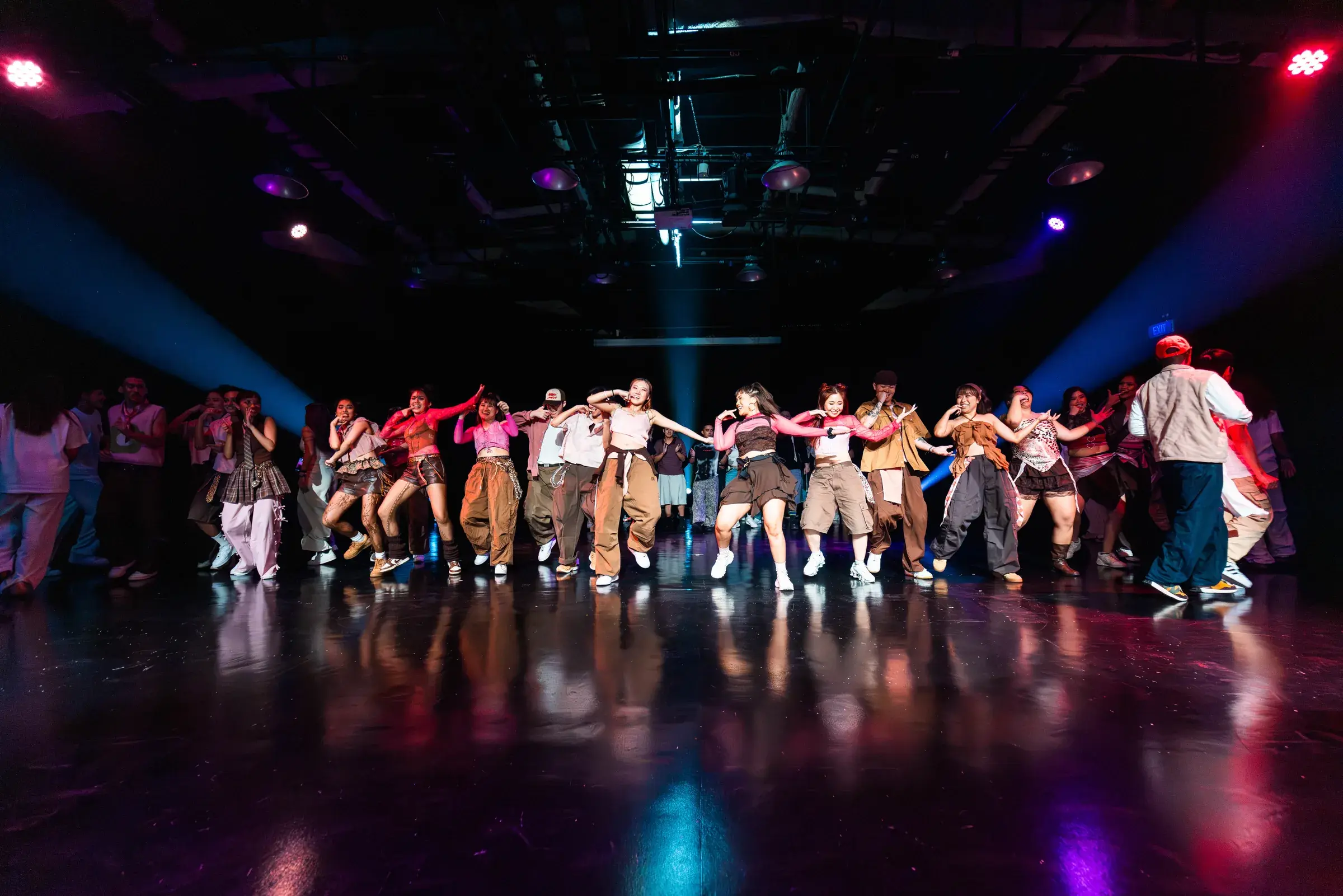 A large group of dancers spread across a dark studio floor mid-routine, arms reaching out in unison under pink and purple stage lighting.
