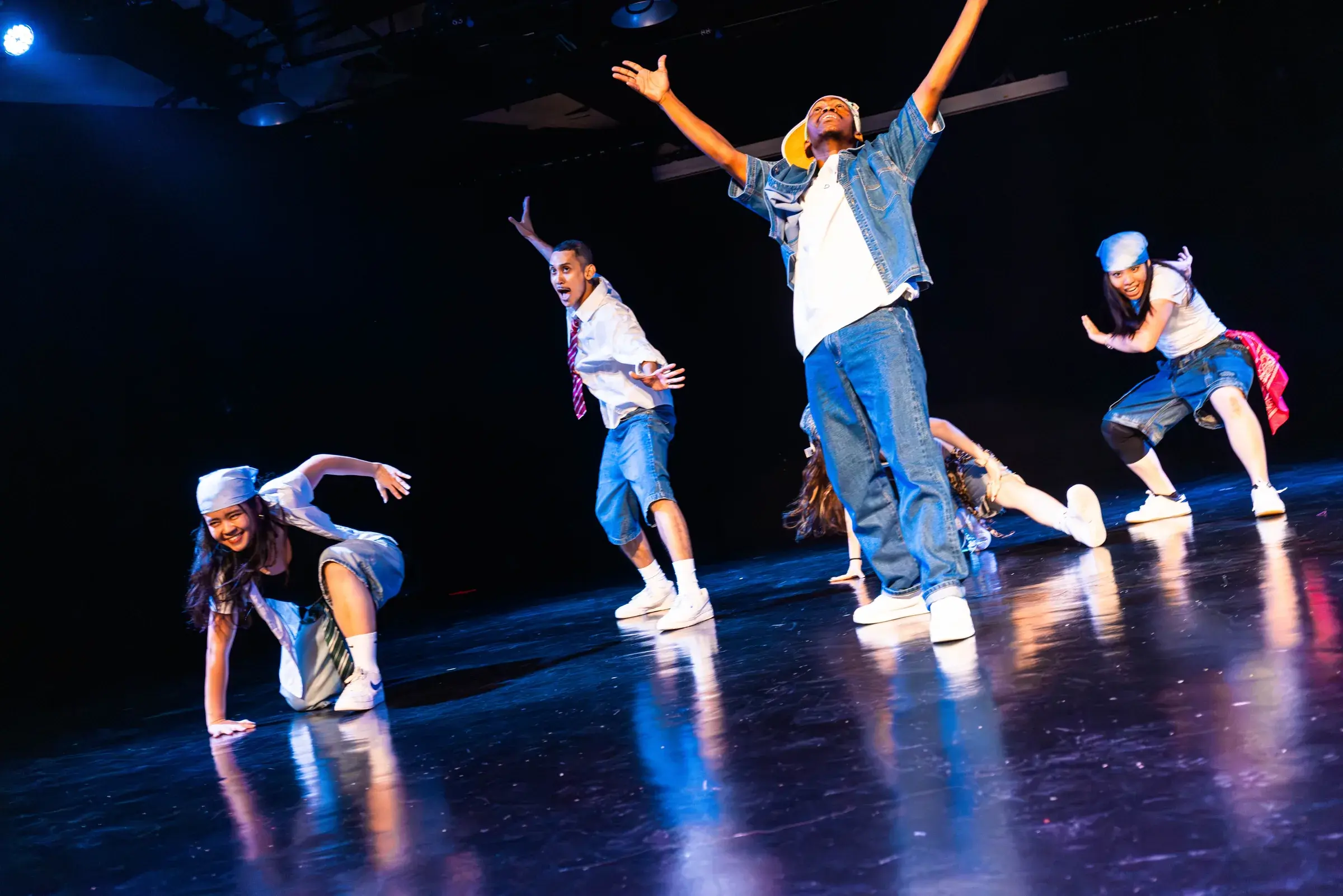A dancer throws both arms open wide, smiling, while three dancers crouch and reach around him.