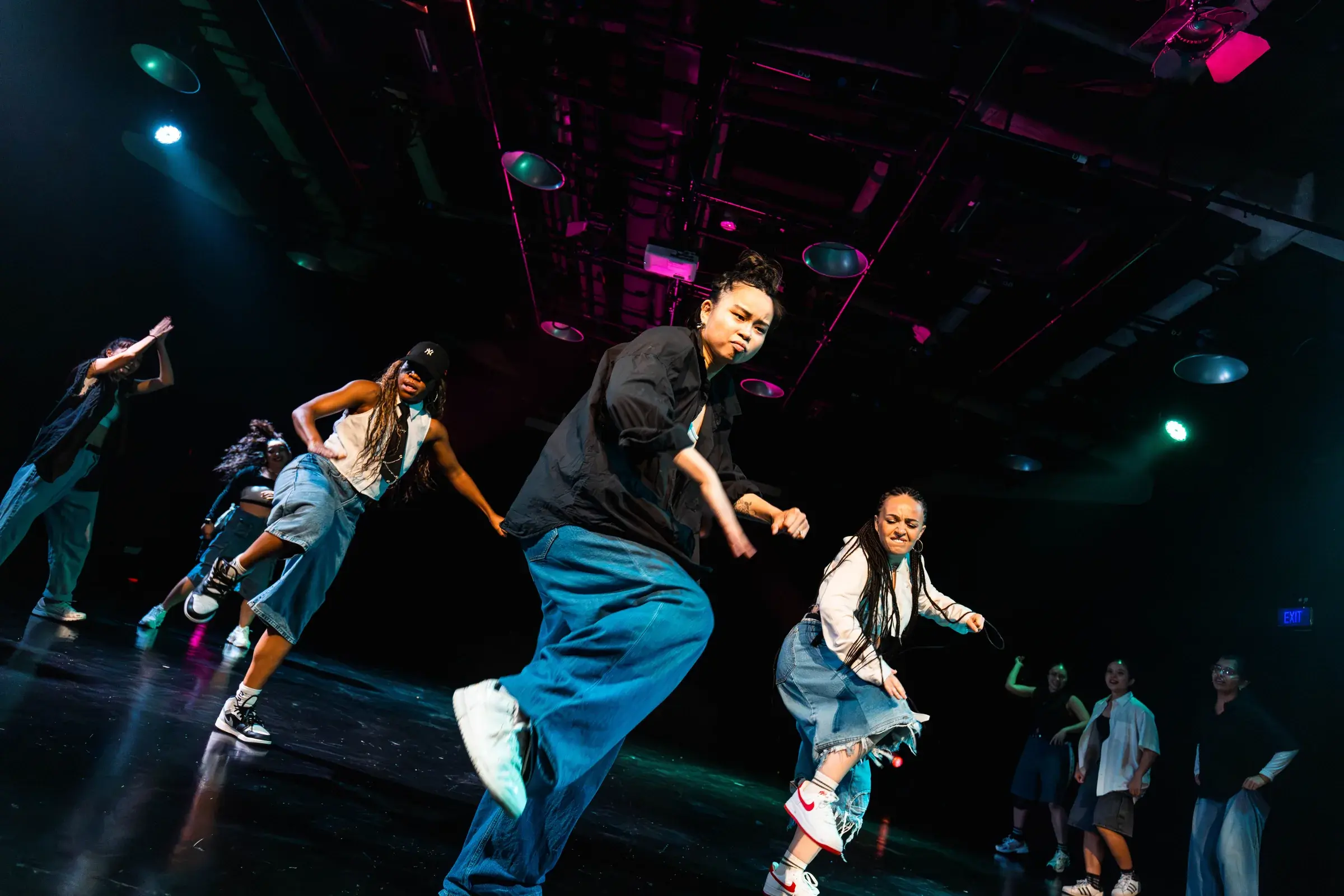 A dancer in teal trousers drops into a deep lunge under blue stage light, other dancers behind.