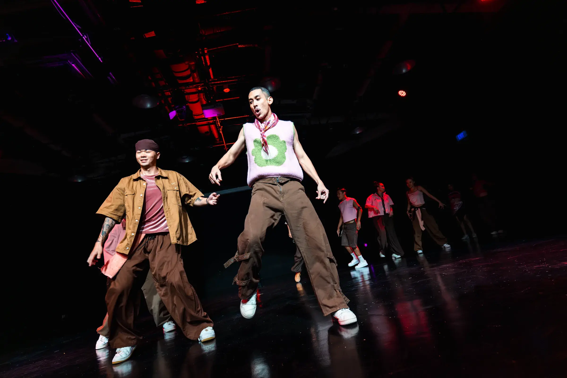 Two dancers in brown and pink stride forward side by side, arms swinging, with a line of dancers behind.