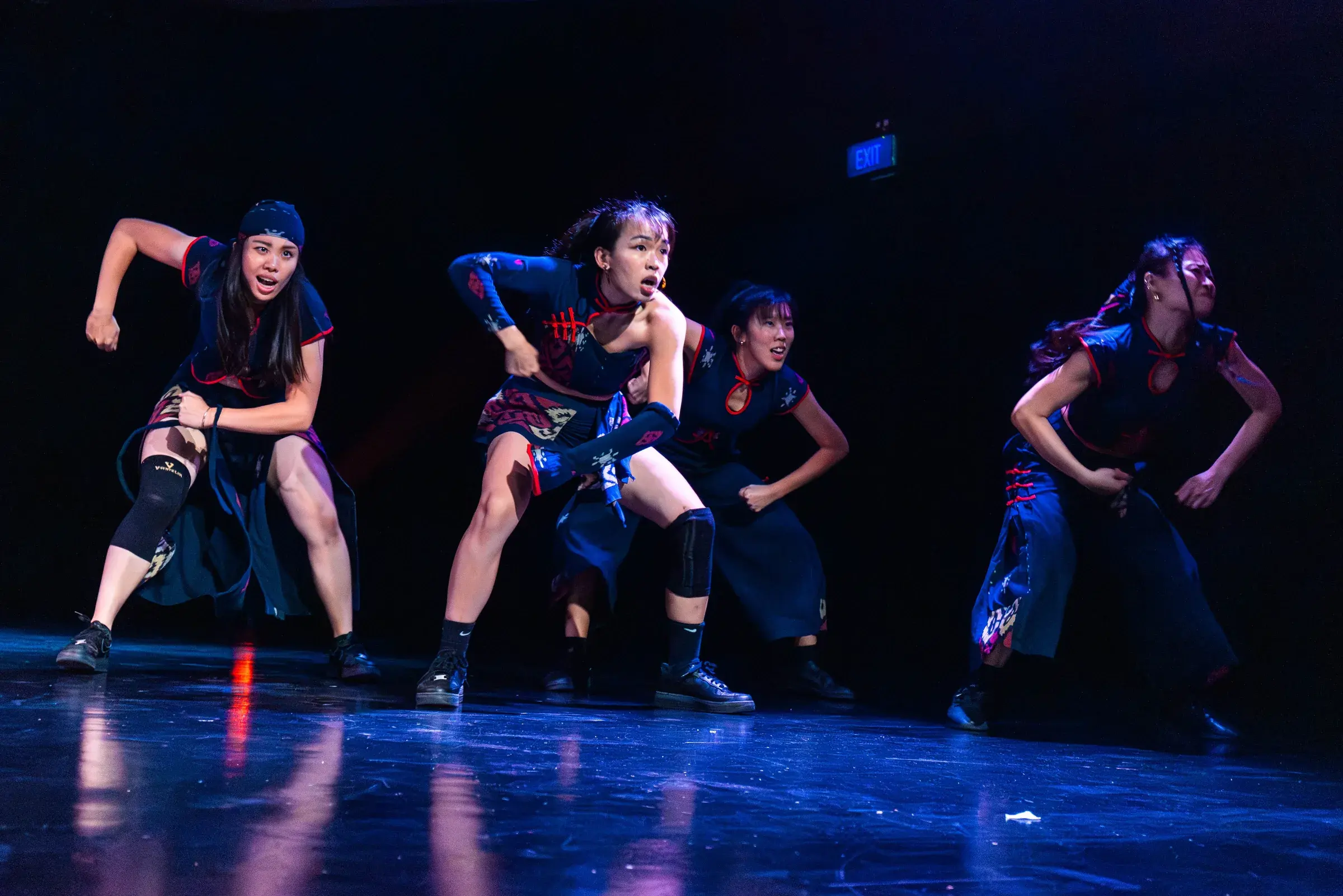 Three dancers in batik-panelled costumes drive forward together under blue light.