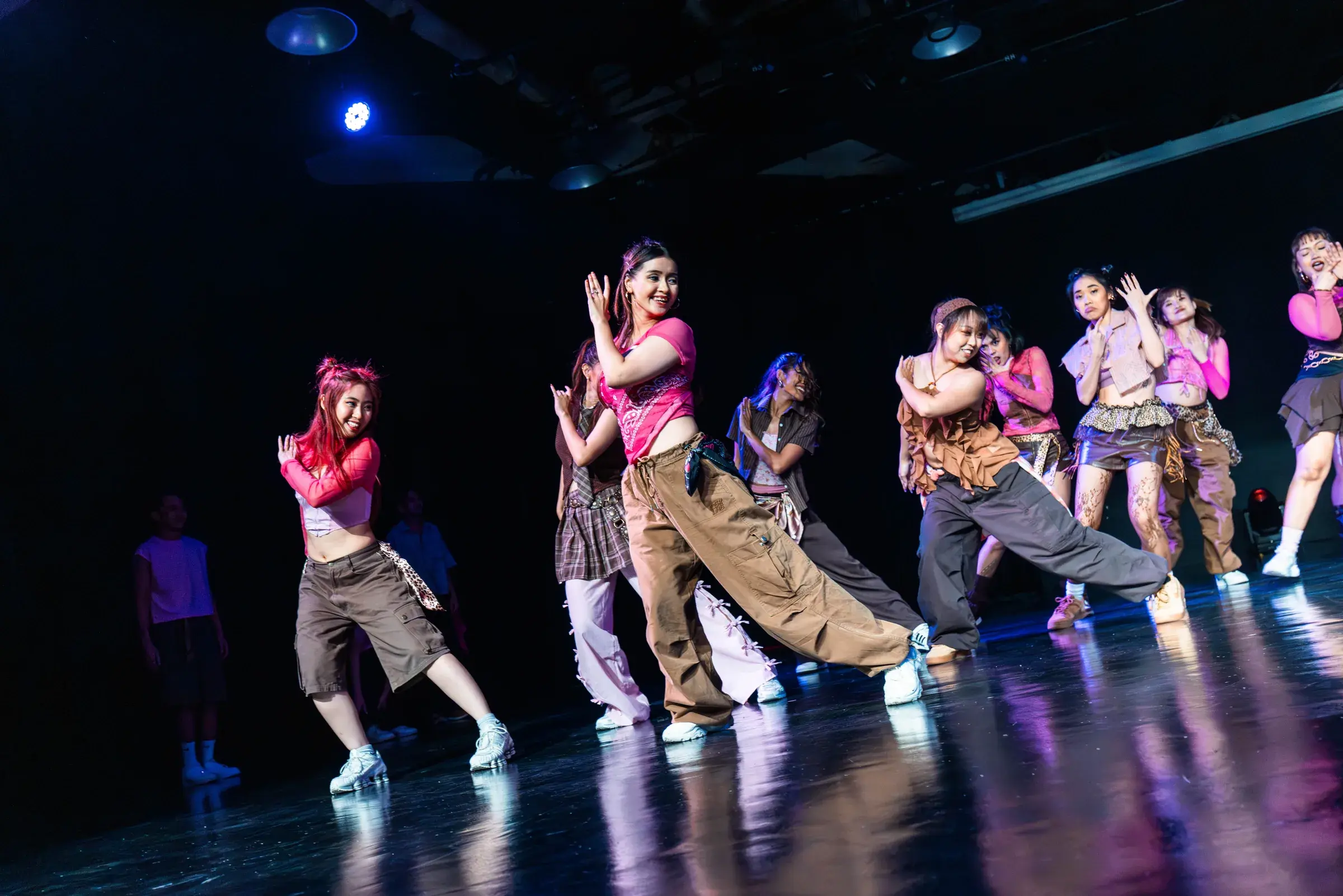 A line of dancers in pink and brown angles across the stage, front dancer reaching one arm out.