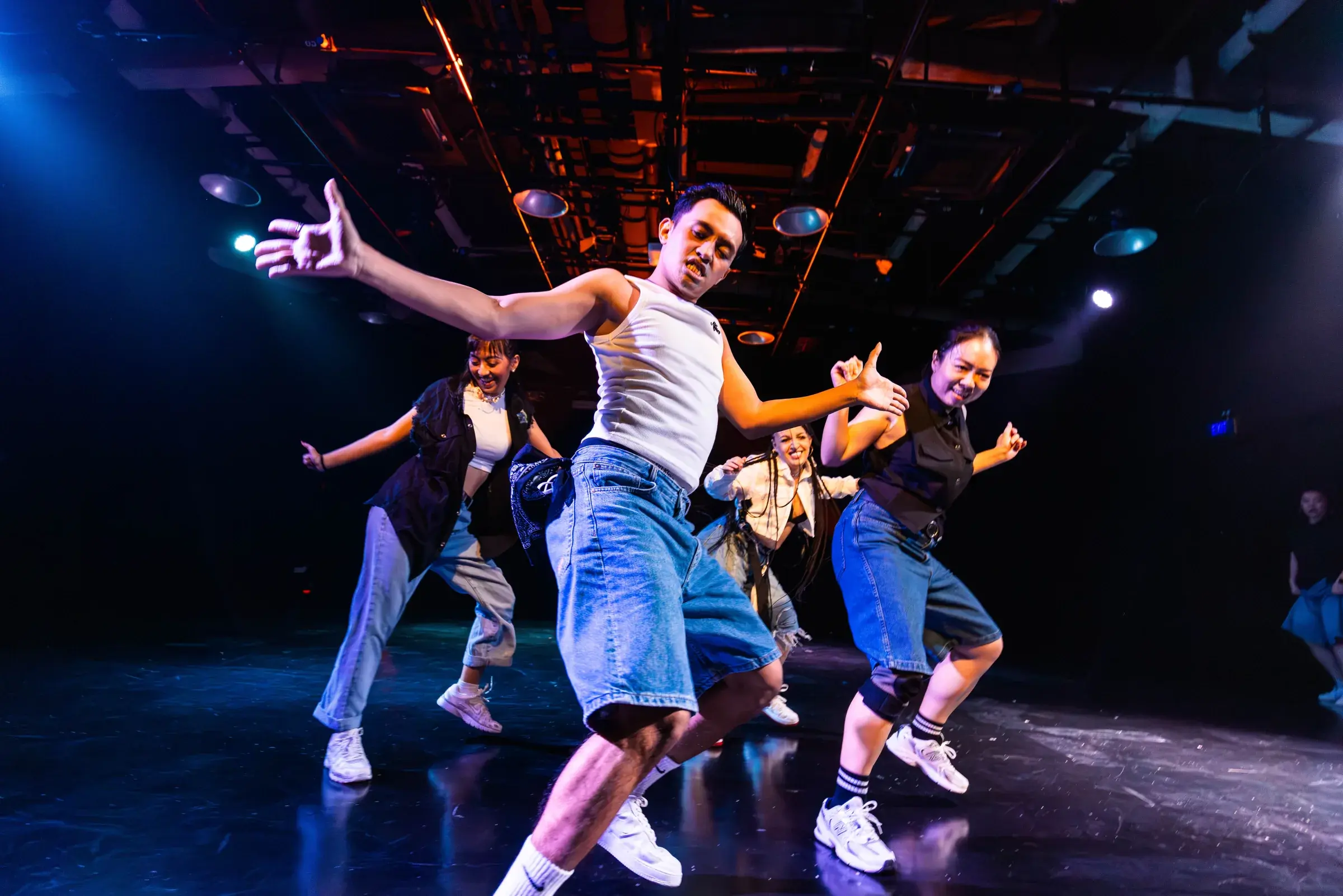 A dancer in a white vest lunges forward with both arms flung wide, four dancers fanned out behind him.
