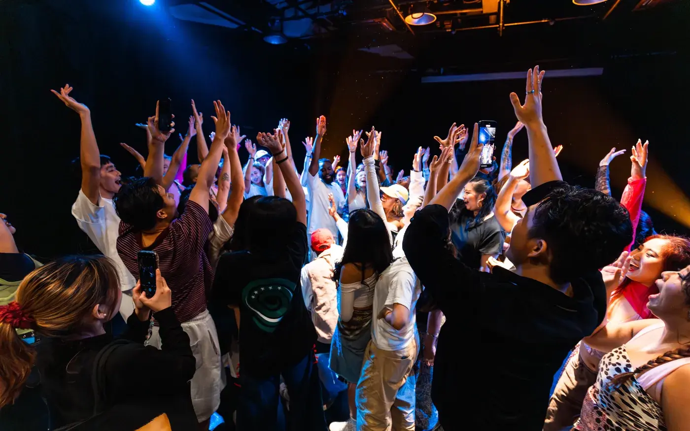 An audience with hands in the air under stage lighting.