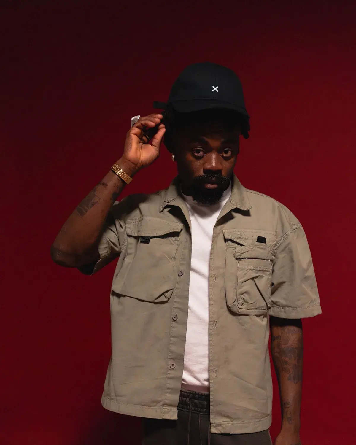 Yemi Osokoya adjusting the brim of his cap, arm tattoos visible, against a deep red studio backdrop.