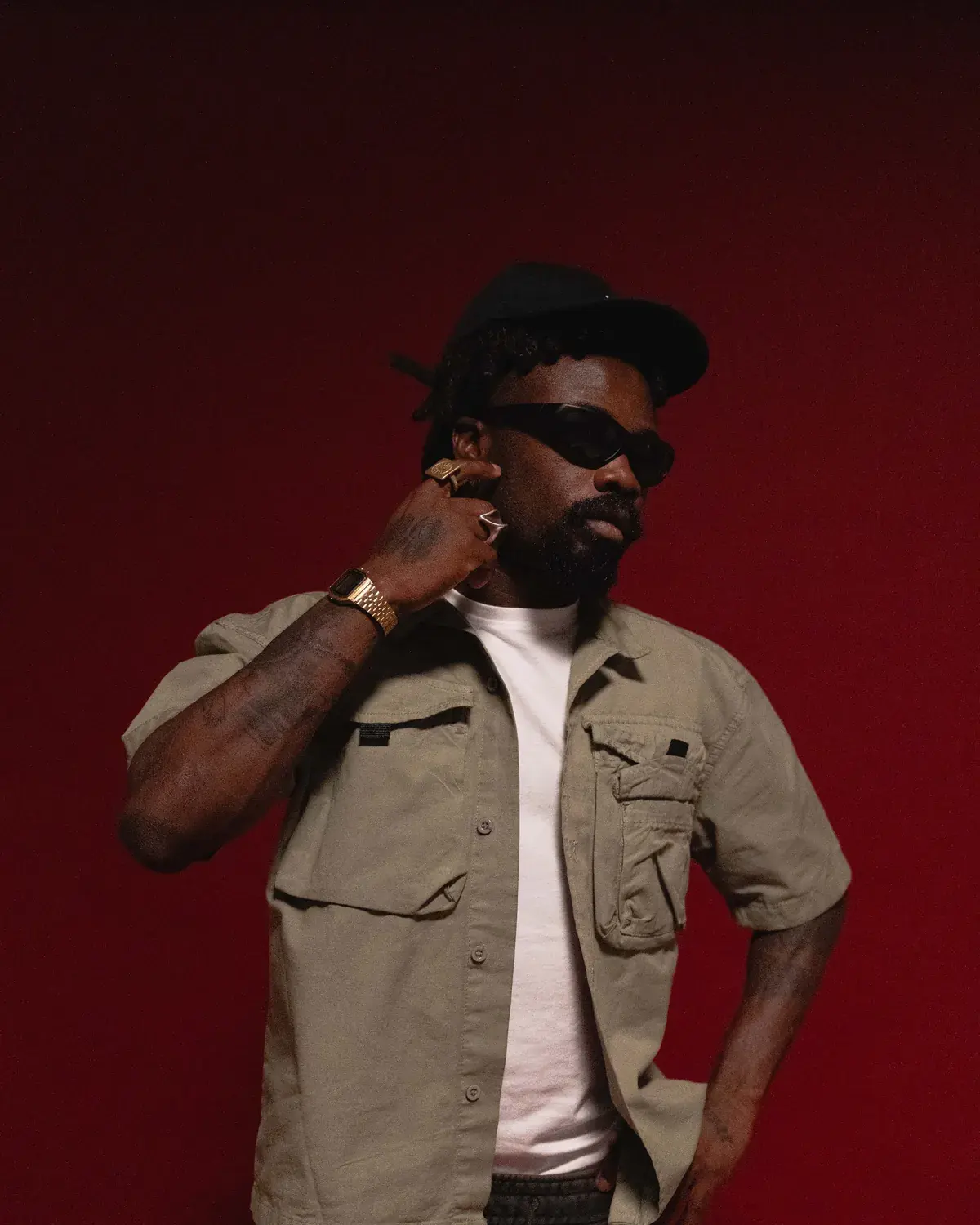 Yemi Osokoya in dark sunglasses with one hand raised to his neck, against a deep red studio backdrop.